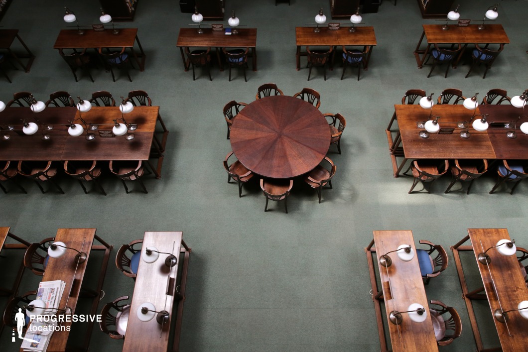  This elegantly arranged library study room features a symmetrical layout of dark wooden tables and chairs, complemented by vintage-style reading lamps with frosted glass shades. A central round table provides a communal study space, contrasting with the traditional long tables that encourage focused individual work. The deep green carpeted floor enhances the warm and scholarly atmosphere, harmonizing with the rich wood tones. The overhead perspective captures the structured arrangement, emphasizing balance and order. This refined setting is ideal for academic, historical, or institutional-themed productions requiring a classic intellectual ambiance.