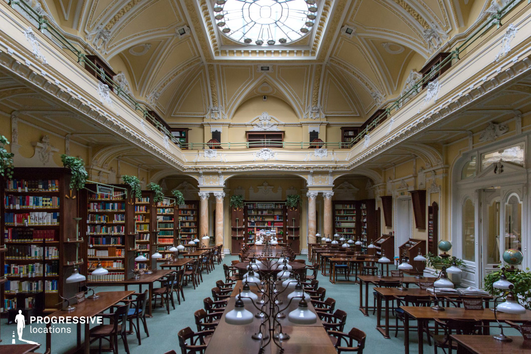 This exquisite library reading room blends classical elegance with a bright, airy ambiance. The high, vaulted ceiling, adorned with intricate white stucco detailing, is crowned by a grand skylight that bathes the space in natural light. Warm pastel tones and decorative molding embellish the walls, while polished wooden bookshelves line the perimeter, filled with neatly arranged volumes. A mezzanine level with an ornate railing adds depth and grandeur to the space. Rows of dark wooden reading tables, illuminated by vintage-style lamps, create an inviting atmosphere for study and research. Lush green plants add a touch of vibrancy, contrasting beautifully with the historic architecture. This location is ideal for period dramas, academic settings, and productions that require a refined yet luminous backdrop.