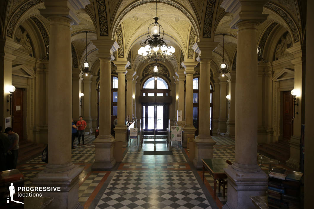 This majestic entrance hall exemplifies classical architecture with its grand vaulted ceilings, intricately decorated arches, and a series of imposing stone columns that create a sense of symmetry and grandeur. The checkered marble flooring adds a refined contrast to the soft beige and cream-toned walls, further emphasizing the hall’s stately character. Large wooden doors and tall arched windows allow natural light to filter in, enhancing the space’s elegant atmosphere. Ornate wrought-iron chandeliers and wall-mounted globe lights provide warm, ambient illumination. This location is ideal for historical or academic-themed productions, offering a timeless and sophisticated setting that conveys prestige and intellectual heritage.