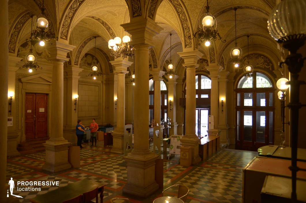 This grand library entrance hall showcases a striking blend of classical architecture and refined elegance. Towering stone columns line the space, supporting a series of intricately decorated vaulted ceilings adorned with delicate ornamental patterns. Large wrought-iron chandeliers and wall-mounted globe lights cast a warm glow, accentuating the depth and texture of the architectural details. The checkered marble floor adds a timeless contrast to the warm beige and cream-toned walls. Tall arched windows and wooden doors invite natural light, enhancing the hall’s stately presence. Ideal for historical, academic, or institutional-themed productions, this space conveys prestige, heritage, and intellectual grandeur.
