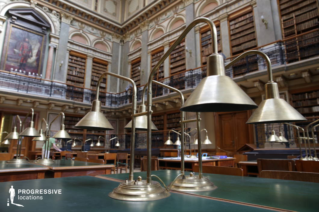 This historic library reading room combines classical elegance with scholarly ambiance. The foreground is dominated by vintage brass reading lamps, their curved necks and wide shades casting a warm, focused glow onto dark green leather-topped desks. The background showcases towering wooden bookshelves, filled with antique volumes and accessible via an ornate mezzanine with wrought iron railings. A large, regal portrait set in a decorative arch adds to the library’s academic prestige. The high, elaborately detailed ceiling enhances the room’s grandeur, while the combination of wood, brass, and deep green hues creates a refined atmosphere ideal for period dramas, literary adaptations, or intellectual-themed productions.