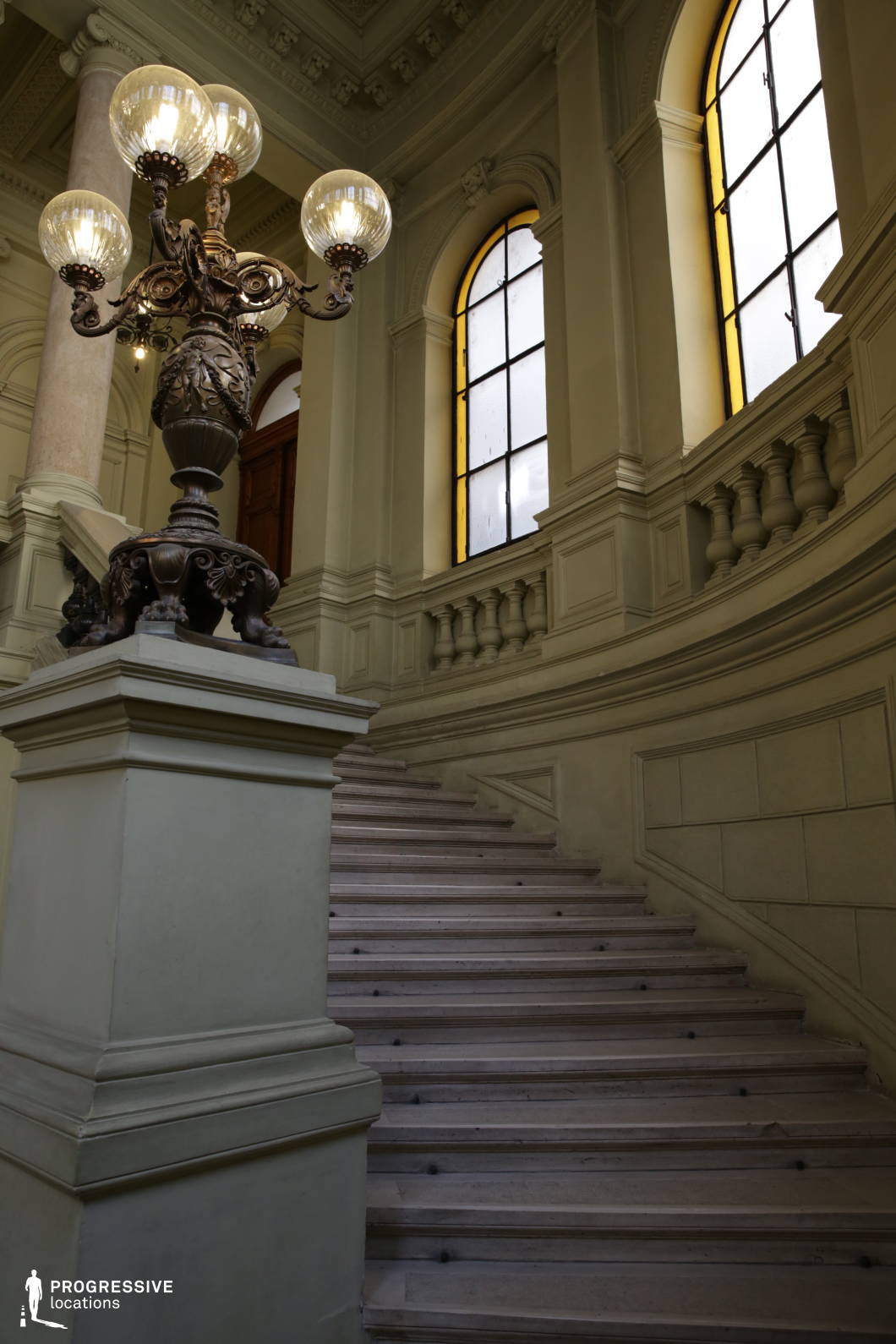 This grand historic staircase exudes classical elegance, featuring wide, gently curving stone steps framed by intricately detailed balustrades. The high, arched windows with soft yellow-tinted glass allow diffused natural light to illuminate the space, enhancing the stately ambiance. A decorative wrought-iron and glass globe lamp post stands prominently, its sculpted base and intricate detailing adding a touch of old-world charm. The refined architectural elements, including fluted columns and decorative moldings, contribute to the staircase’s sophisticated grandeur. This location is ideal for period dramas, historical settings, or productions requiring a regal, atmospheric backdrop.