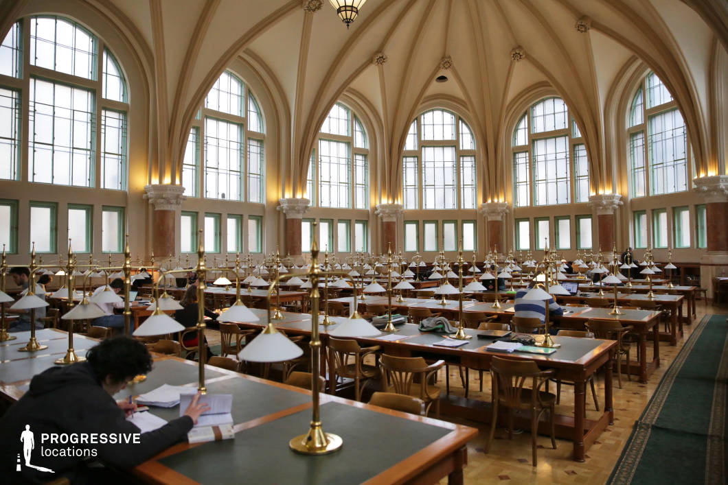 This grand university library reading hall is a stunning example of neo-Gothic architecture, featuring soaring vaulted ceilings supported by elegant ribbed arches. Large arched windows fill the space with natural light, illuminating the symmetrical rows of wooden study tables topped with brass and white-shaded reading lamps. The high ceilings, adorned with intricate decorative elements, enhance the room’s openness and historic charm. The polished wooden flooring and green leather desk surfaces contribute to a refined academic atmosphere. This setting is ideal for scholarly, historical, or period-themed productions, offering a visually striking backdrop that conveys intellectual depth and architectural grandeur.