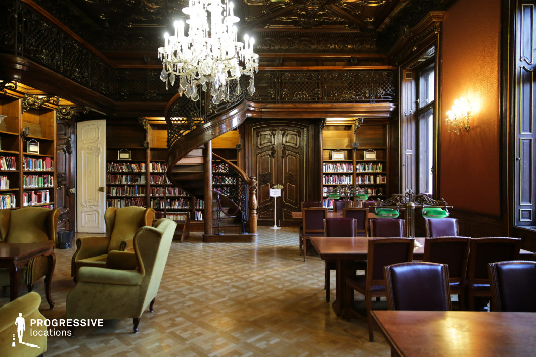 This opulent historic library features an exquisitely carved wooden spiral staircase leading to a mezzanine-level balcony with ornate wrought iron railings. A dazzling crystal chandelier hangs from a dark, intricately detailed ceiling, casting a warm glow over the room. Rich wooden bookshelves filled with colorful volumes line the walls, while classic green banker’s lamps adorn the polished reading tables. Plush vintage armchairs provide a cozy reading area, complementing the parquet flooring and deep red damask wallpaper. This refined setting is ideal for period dramas, academic or literary-themed productions, and scenes requiring a stately, aristocratic ambiance.