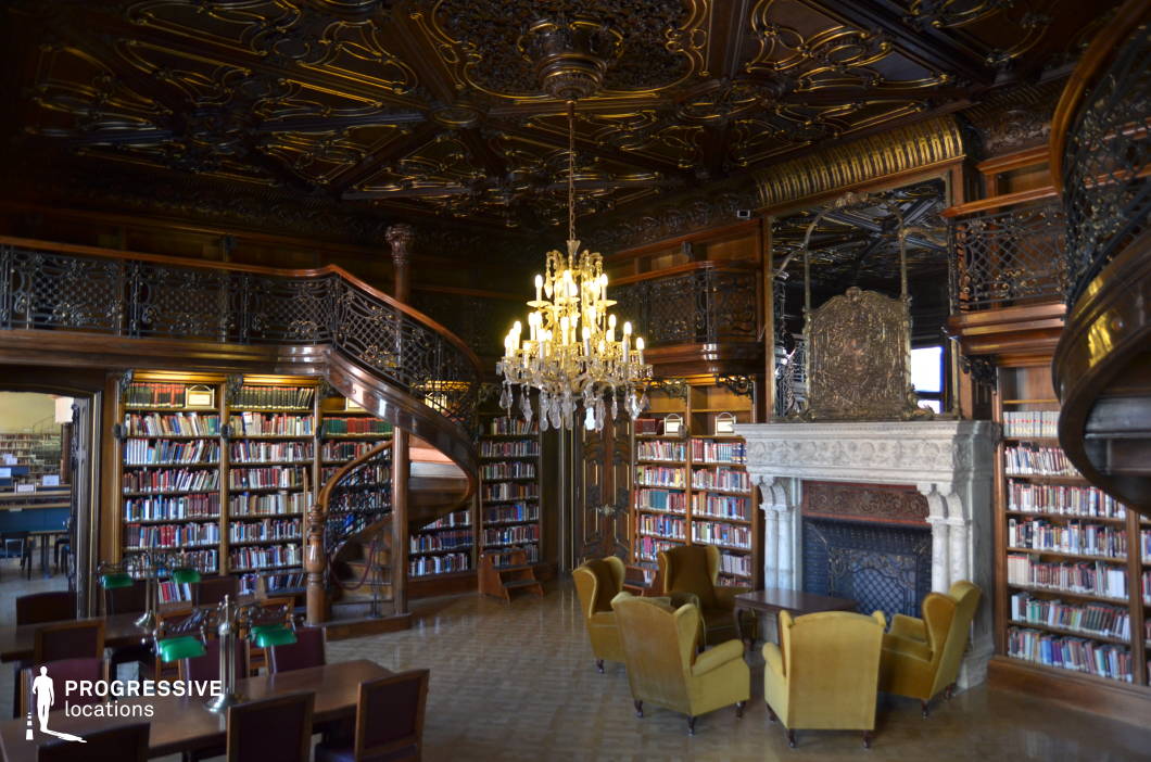 This opulent historic library boasts intricate woodwork, an elaborately carved ceiling, and a grand chandelier that casts a warm, atmospheric glow over the space. Rich wooden bookshelves filled with colorful volumes line the walls, accessible via elegant spiral staircases leading to a mezzanine-level balcony with ornate wrought iron railings. A stately stone fireplace, adorned with an intricately detailed relief, anchors the room, enhancing its aristocratic charm. The plush, vintage-style seating in mustard-colored upholstery creates an inviting reading area, contrasting with the polished parquet flooring. This visually striking location is ideal for period dramas, classic literature adaptations, or intellectual-themed productions, evoking an air of sophistication and exclusivity.