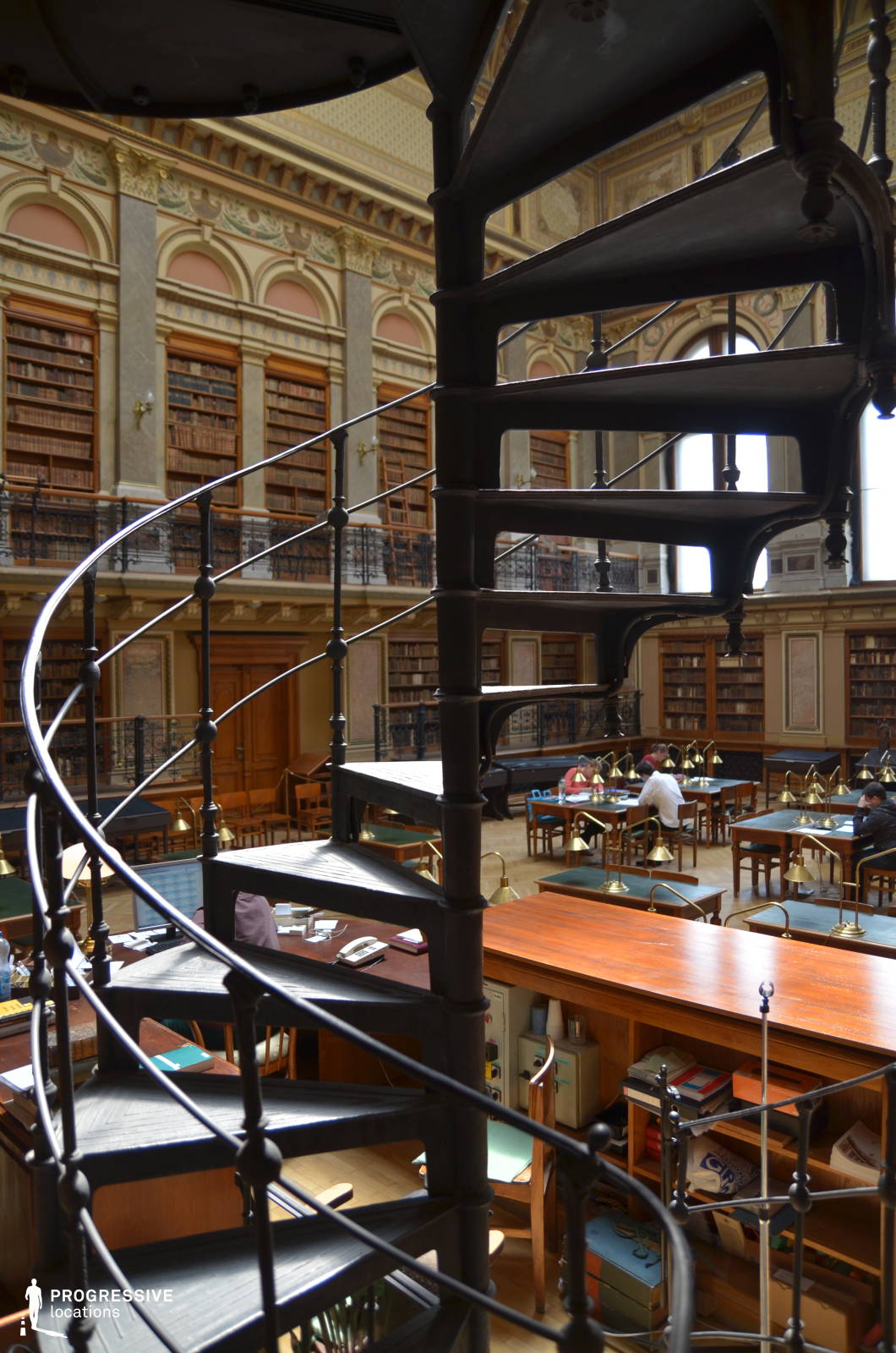 This striking spiral staircase, crafted from dark wrought iron, serves as a stunning architectural feature within a grand historic library. Its delicate curves and intricate railing design contrast beautifully with the soaring bookshelves and classical decor in the background. The room is bathed in warm natural light, highlighting the ornate detailing on the walls, the mezzanine’s decorative ironwork, and the rich wooden furnishings. Rows of brass reading lamps line the study tables, reinforcing the scholarly atmosphere. This visually compelling setting is ideal for period dramas, literary-themed productions, or scenes requiring an air of intellectual grandeur and historical refinement.