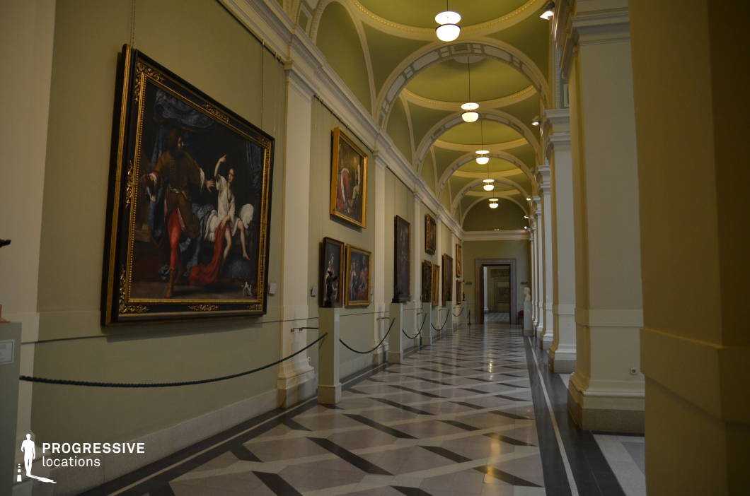 This refined museum gallery features a long, arched corridor with soft green vaulted ceilings, accented by white decorative moldings that add a touch of classical elegance. The walls are adorned with large, ornately framed oil paintings, creating a rich visual narrative along the length of the space. The polished marble floor, designed with a geometric black and white pattern, enhances the sense of depth and grandeur. Subtle lighting from hanging globe fixtures casts a warm glow, emphasizing the intricate architectural details and artistic displays. The combination of symmetry, depth, and classic aesthetics makes this gallery an ideal setting for period dramas, historical documentaries, or high-end fashion photography.