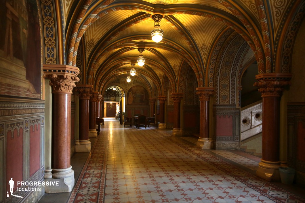 This richly decorated Gothic Revival hallway features a series of ribbed, pointed arches supported by polished red marble columns with intricately carved capitals, creating a visually immersive architectural space. The ceiling and walls are adorned with detailed geometric and floral motifs in warm, earthy tones, enhancing the historic and regal ambiance. A patterned tile floor runs the length of the corridor, bordered by ornamental designs that complement the surrounding decorative elements. Hanging lantern-style chandeliers cast a soft, golden glow, emphasizing the depth and symmetry of the passage. The interplay of light, color, and texture makes this location ideal for historical, fantasy, or period settings requiring an opulent and atmospheric backdrop.