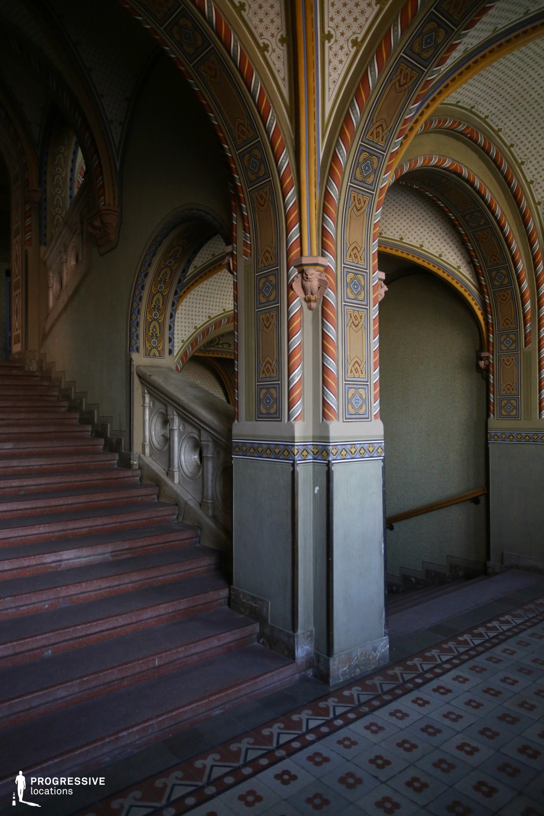 This grand staircase is framed by richly decorated Gothic-style arches, adorned with intricate geometric and floral patterns in earthy tones of red, gold, and blue. The columns and vaulted arches feature painted detailing with repeating motifs, while sculpted stone corbels add a sense of craftsmanship and historic authenticity. The staircase itself is made of polished stone, with an elegantly carved balustrade that complements the surrounding architecture. The patterned tile flooring enhances the sense of refinement, while the interplay of light and shadow emphasizes the depth and structure of the space. This visually striking setting is ideal for historical, fantasy, or period productions requiring a richly textured and atmospheric backdrop.
