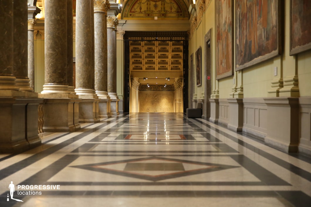 This opulent classical hallway features towering marble columns with gilded capitals, a polished geometric-patterned marble floor, and high ceilings adorned with intricate gold detailing. The symmetrical colonnade creates a sense of depth and grandeur, while the walls are lined with large historical paintings in ornate frames, enhancing the refined and museum-like ambiance. At the far end, an elaborately decorated entrance with carved embellishments and warm lighting draws the eye, adding to the architectural splendor. The fusion of symmetry, luxurious materials, and historic charm makes this setting ideal for period dramas, cultural documentaries, or elegant fashion shoots.