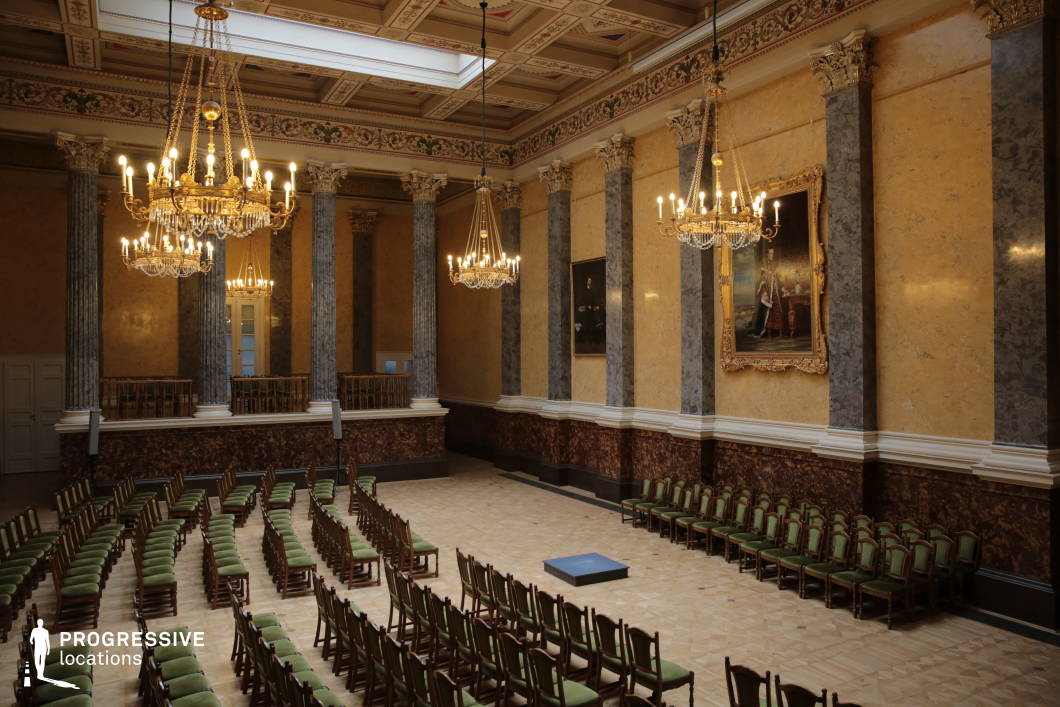 This grand neoclassical assembly hall features towering marble columns, gilded chandeliers, and intricately decorated walls, exuding an atmosphere of refinement and prestige. The high, coffered ceiling is adorned with floral and geometric motifs, while large gold-framed portraits enhance the historic character of the space. The symmetrical arrangement of green-upholstered wooden chairs and a central speaking platform suggest a formal setting, ideal for ceremonies, lectures, or high-profile gatherings. Warm ambient lighting from the chandeliers complements the rich color palette of golden walls, dark marble paneling, and polished parquet flooring, making this location perfect for period dramas, historical reenactments, or sophisticated cultural events.