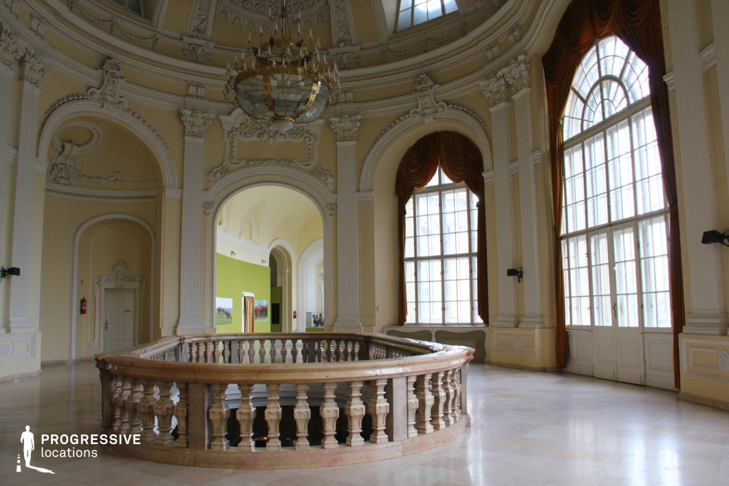 This elegant Baroque hall features soaring ceilings adorned with intricate stucco moldings, grand arched windows draped in gold-hued curtains, and a stunning central chandelier that enhances the space’s opulent character. The soft cream and ivory tones of the walls, complemented by classical pilasters and sculpted reliefs, create a refined and airy ambiance. A curved stone balustrade encircles an open mezzanine level, adding depth and architectural sophistication. The polished marble flooring reflects the natural light streaming in through the expansive windows, further accentuating the room’s luminosity. This location is ideal for historical productions, grand interior scenes, or luxury event settings requiring a stately and regal atmosphere.
