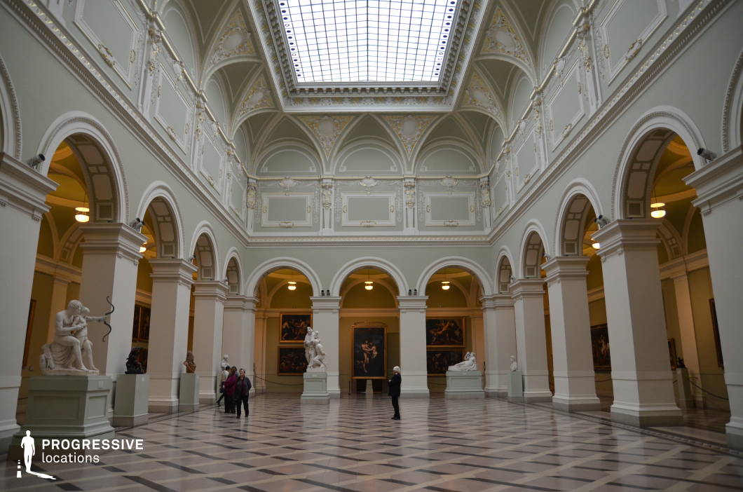 This grand neoclassical museum hall features towering arched openings, intricately decorated white and pastel-green walls, and a coffered ceiling with gold embellishments, all illuminated by a central skylight that floods the space with natural light. The symmetrical design, supported by massive columns, enhances the sense of balance and elegance, while the polished marble floor with geometric patterns reflects the classical architectural style. Marble sculptures are strategically placed throughout the hall, adding a timeless artistic element, while large oil paintings adorn the surrounding gallery walls. The combination of scale, refined detailing, and luminous atmosphere makes this setting ideal for historical productions, museum exhibitions, or sophisticated editorial shoots.