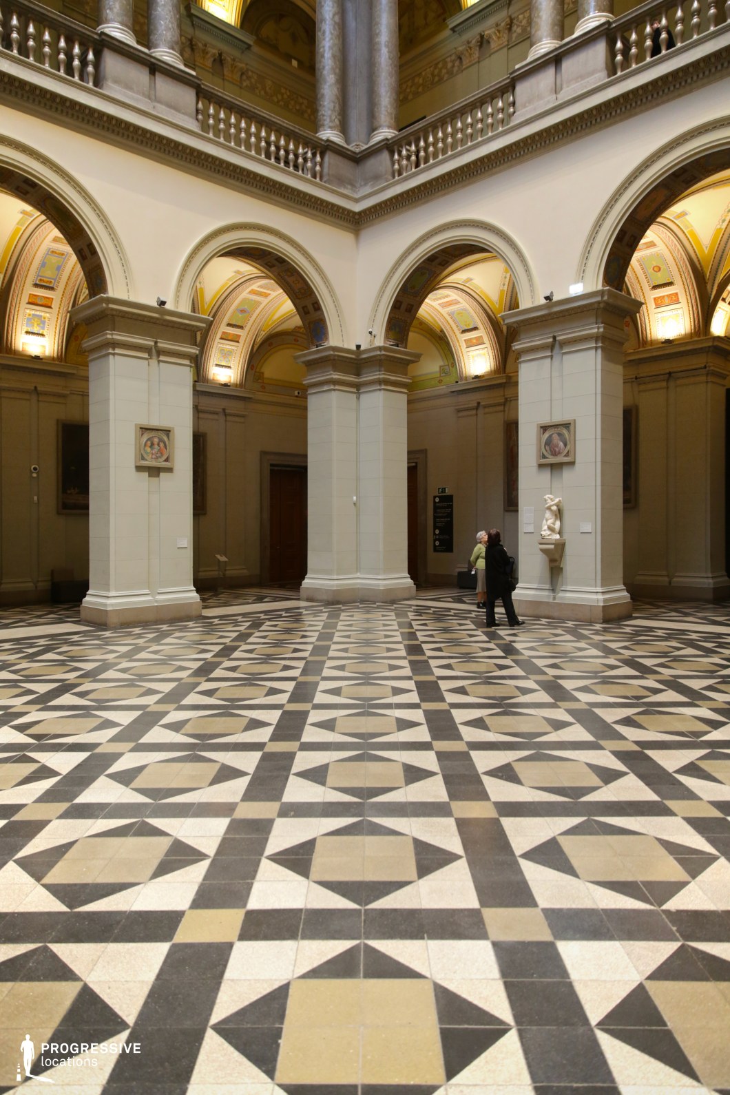 This grand neoclassical hall features towering arched openings, massive stone columns, and an intricately patterned geometric floor that enhances the depth and symmetry of the space. The lower level is defined by smooth, cream-colored walls accented with sculptural reliefs and classical busts, while the upper gallery is framed by balustrades and marble-clad columns supporting the structure. The vaulted ceilings are adorned with richly painted frescoes in warm golden hues, adding a sense of historic opulence. Soft, ambient lighting highlights the architectural details, making this space ideal for period films, museum scenes, or high-end editorial productions requiring a regal and stately setting.