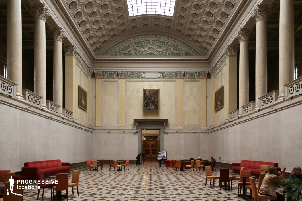 This stately neoclassical hall features soaring marble columns, a coffered vaulted ceiling, and grand arched details that emphasize its scale and elegance. The muted cream and green palette, complemented by delicate ornamental friezes and gold-accented paintings, enhances the historic and refined atmosphere. The floor is adorned with a geometric black-and-white mosaic pattern, creating a sense of depth and symmetry. The space is furnished with contemporary chairs and red sofas, blending classical architecture with modern functionality. Soft natural light filters in through a skylight, illuminating the intricate details and architectural harmony, making this hall an ideal location for cultural events, historical productions, or sophisticated editorial settings.