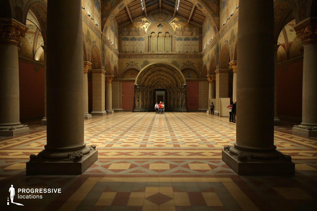 This majestic Romanesque hall features towering stone columns, intricate gold-accented capitals, and a richly patterned tile floor that enhances its historical grandeur. The high, vaulted ceiling is adorned with elaborate frescoes depicting classical motifs, while arched walkways and sculptural details contribute to the space’s sense of depth and symmetry. At the far end, a grand semicircular apse with an intricately carved portal serves as the focal point, framed by warm, ambient lighting. The interplay of earthy tones, gilded embellishments, and medieval-inspired architecture makes this location ideal for historical productions, period dramas, or immersive museum settings requiring a dramatic and atmospheric backdrop.