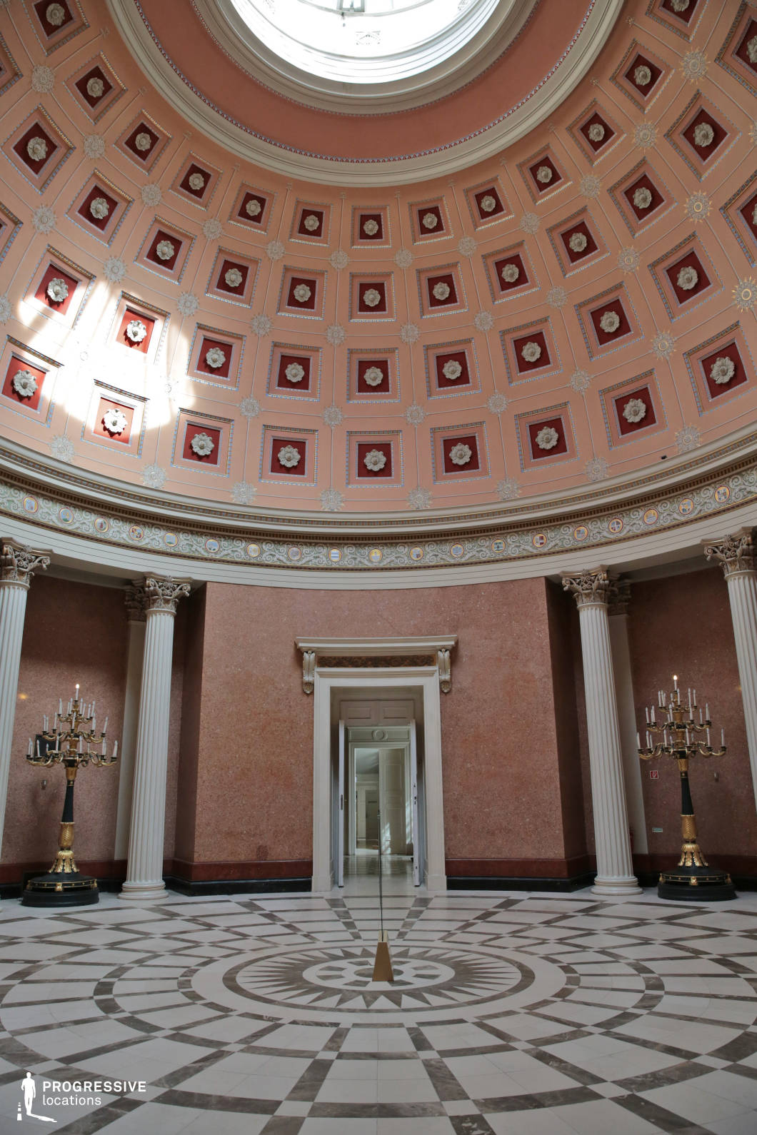 This striking neoclassical rotunda features a vast domed ceiling adorned with square coffers, each decorated with floral reliefs in red and gold tones, creating a sense of depth and symmetry. Towering Corinthian columns frame the entrance, leading to a spacious, circular chamber with polished marble walls and a geometric-patterned floor that radiates outward from a central focal point. Elegant gilded candelabras add a regal touch, while the soft natural light filtering through the skylight highlights the intricate architectural details. The harmonious balance of classical proportions, refined ornamentation, and a grand spatial composition makes this an ideal location for historical productions, museum settings, or prestigious formal events.