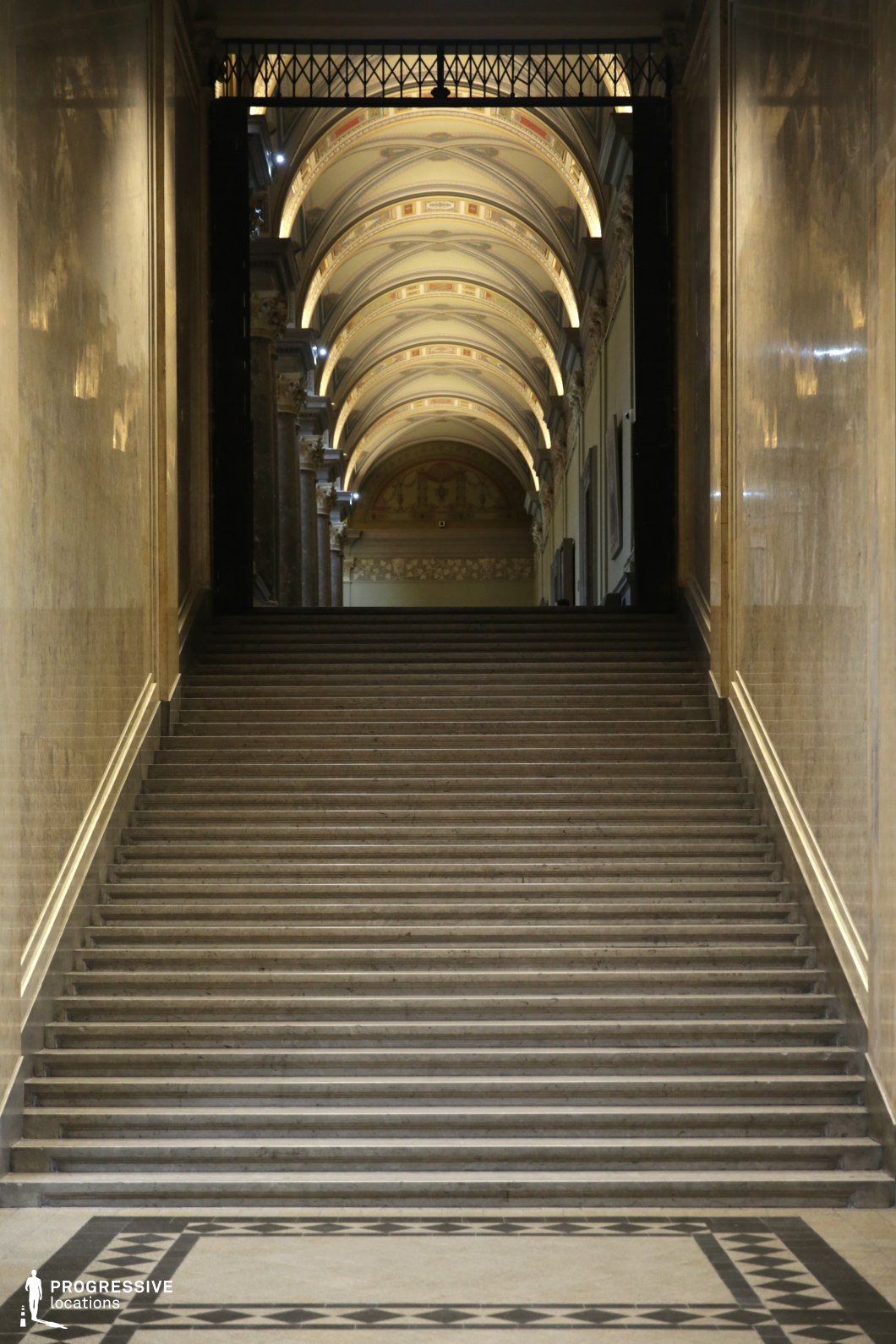 This imposing neoclassical staircase is framed by towering marble walls and leads to a long, vaulted corridor adorned with intricate frescoes and softly illuminated archways. The wide, symmetrical steps are made of polished stone, creating a sense of grandeur and formality, while the geometric patterned flooring at the base adds a refined architectural detail. The ceiling’s rhythmic repetition of arches and soft lighting enhances the depth and perspective, drawing the eye towards the upper level. The combination of classical elegance, dramatic lighting, and imposing scale makes this location ideal for historical productions, formal institutional settings, or cinematic moments requiring a majestic and atmospheric backdrop.