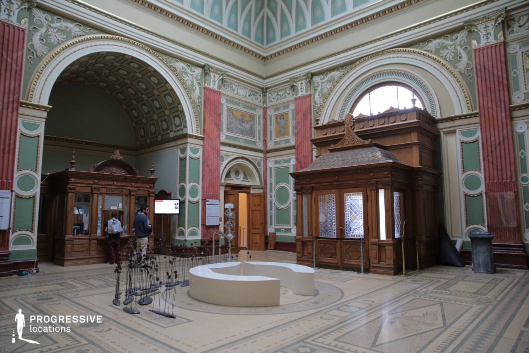 This grand neoclassical entrance hall features intricately detailed pastel green walls with gold-accented stucco moldings, large arched windows, and decorative red marble pilasters that enhance the classical aesthetic. The high coffered ceiling and ornamental frescoes add a sense of depth and historic refinement. Dominating the space are two finely crafted wooden ticket booths with elaborate detailing and lattice windows, blending seamlessly with the surrounding architecture. The patterned mosaic floor complements the hall’s sophisticated ambiance, while contemporary artistic elements introduce a modern contrast. This striking fusion of classical elegance and functional design makes the space ideal for museum settings, cultural venues, or historical period productions.