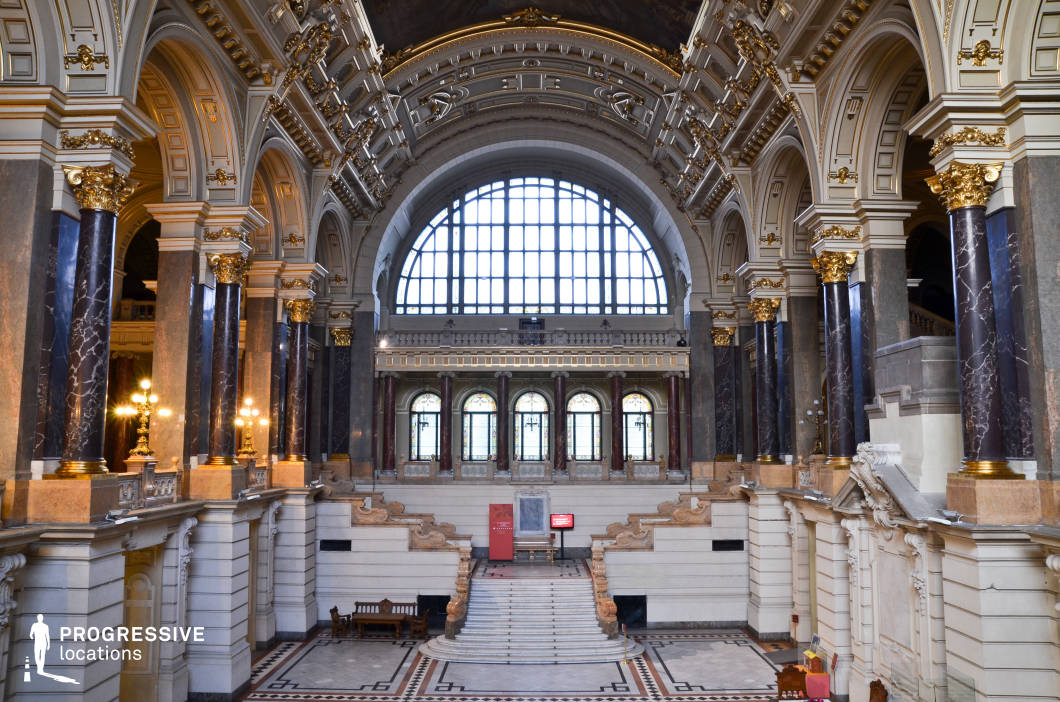 This opulent Baroque Revival hall features towering black marble columns adorned with gilded Corinthian capitals, a coffered ceiling with elaborate gold detailing, and grand arched openings that enhance the sense of scale and refinement. A sweeping staircase with intricately carved balustrades leads to an elevated platform framed by stained-glass windows and an expansive arched skylight, flooding the space with natural light. The floor showcases a geometric mosaic pattern that complements the symmetrical architecture, while classic furnishings and decorative wall sculptures add to the hall’s regal ambiance. The combination of monumental scale, rich ornamentation, and dramatic lighting makes this location ideal for historical dramas, prestigious events, or high-end fashion editorials requiring a majestic setting.