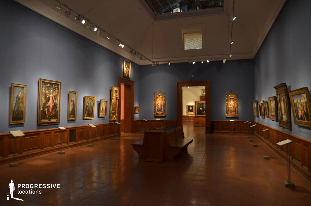This intimate and elegantly curated art gallery features deep blue walls, warm wooden paneling, and a terracotta-tiled floor that enhances the historic ambiance. The space is illuminated by adjustable spotlights, which highlight the collection of religious and Renaissance-style paintings framed in gilded and carved wood. A central wooden bench provides a resting point, while an open doorway leads to another exhibition room, creating a sense of continuity. The combination of subdued lighting, classic architectural detailing, and rich color contrasts makes this setting ideal for historical documentaries, refined cultural presentations, or period-inspired artistic productions.
