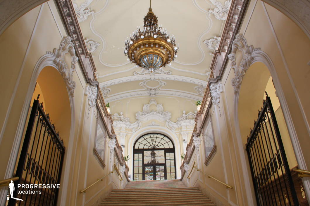 This magnificent Baroque-style staircase exudes elegance with its sweeping marble steps, intricately carved stucco moldings, and soft cream-colored walls adorned with sculptural reliefs. An opulent chandelier with gold and blue accents hangs from the elaborately decorated ceiling, casting a warm glow over the space. The wrought-iron gates with gold handrails add a regal touch, while the arched glass doorway at the top enhances the grandeur by providing a glimpse into the hall beyond. The harmonious combination of classical ornamentation, refined architectural elements, and luxurious materials makes this location ideal for period dramas, historical productions, or high-end editorial photography.