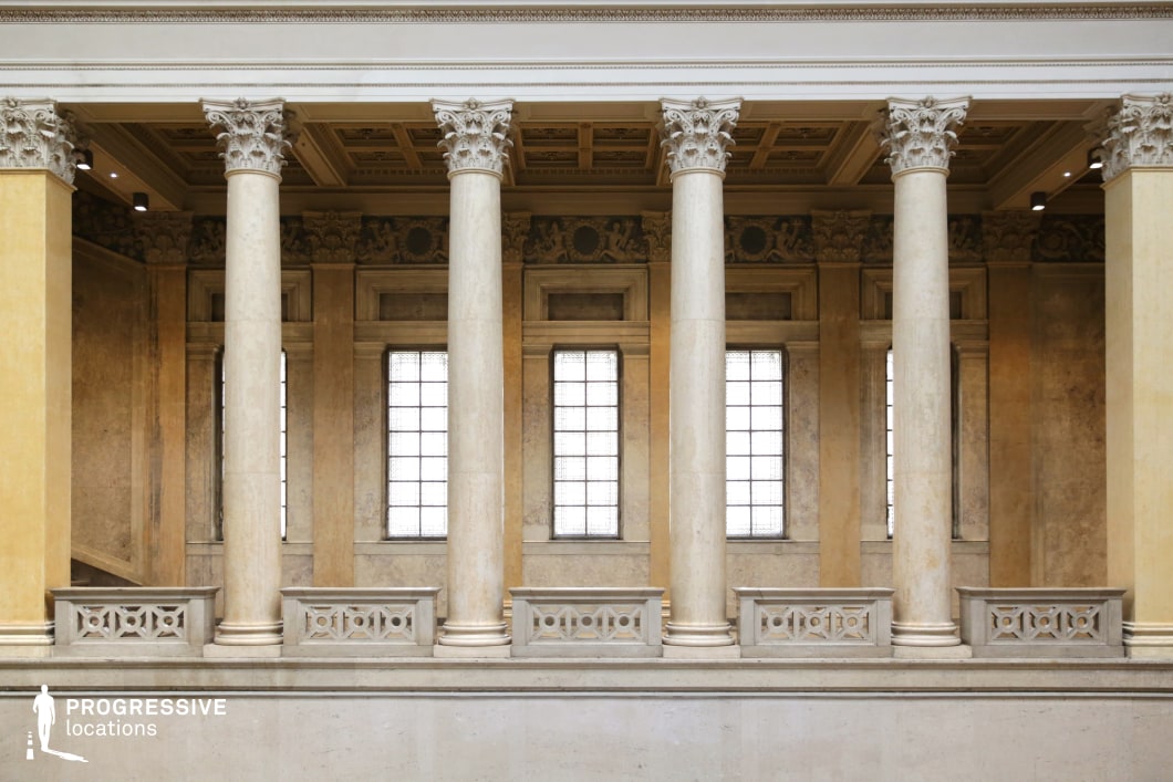 This stately neoclassical colonnade features towering Corinthian columns made of smooth white marble, supporting an intricately coffered ceiling that enhances the architectural grandeur. The symmetrical arrangement of columns and balustrades creates a rhythmic visual flow, while the textured beige walls and tall, frosted windows add depth and elegance. Delicate sculptural reliefs and ornamental friezes adorn the upper sections, reinforcing the refined classical aesthetic. The soft, diffused natural light filtering through the windows highlights the intricate detailing, making this location ideal for historical settings, formal institutional scenes, or elegant architectural photography.