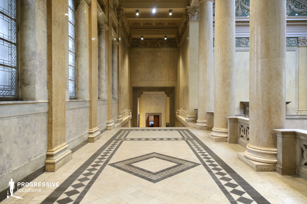 This stately neoclassical corridor is defined by towering marble columns, intricate coffered ceilings, and elegant geometric-patterned flooring that enhances its sense of symmetry and grandeur. The walls are clad in polished cream-colored marble, reflecting soft ambient lighting, while tall frosted windows framed by decorative moldings allow diffused natural light to illuminate the space. Ornate balustrades and sculptural friezes add a refined classical touch, reinforcing the monumental scale of the architecture. The seamless blend of sophistication, depth, and symmetry makes this location ideal for historical settings, prestigious institutions, or cinematic productions requiring a majestic and formal atmosphere.
