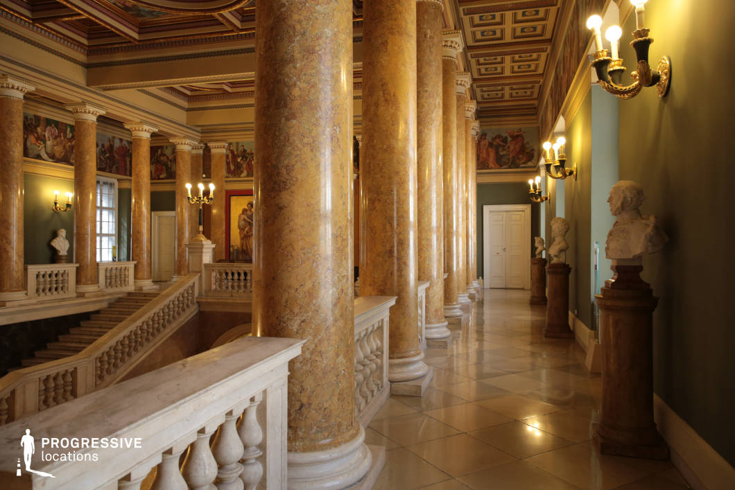 This opulent neoclassical interior features towering golden marble columns, an ornate coffered ceiling with intricate detailing, and a grand staircase framed by sculpted balustrades. The walls are adorned with classical frescoes and gilded wall sconces, casting a warm ambient glow that enhances the regal atmosphere. White marble busts on pedestals line the hallway, adding to the sense of historical grandeur, while the polished tiled floor reflects the soft lighting, amplifying the space’s elegance. The symmetrical arrangement, rich textures, and classical artistry make this location ideal for period dramas, historical documentaries, or refined cultural settings requiring a prestigious and majestic backdrop.