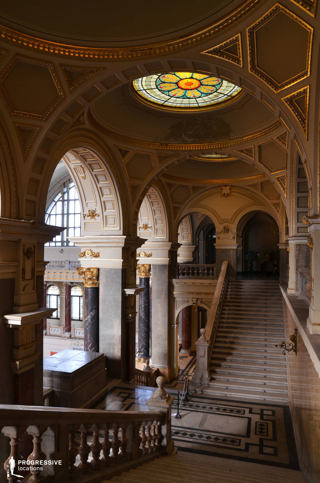 This magnificent Baroque Revival staircase features soaring arches, intricately gilded moldings, and towering marble columns that exude grandeur and sophistication. A striking stained-glass skylight with vibrant floral motifs filters natural light, casting a warm glow over the coffered ceiling and elegant architectural details. The wide stone staircase, framed by sculpted balustrades, leads to a series of opulent archways, enhancing the sense of depth and scale. Polished marble flooring with geometric inlays further accentuates the stately ambiance, making this location ideal for period dramas, historical settings, or luxury-themed productions requiring a regal and visually immersive backdrop.