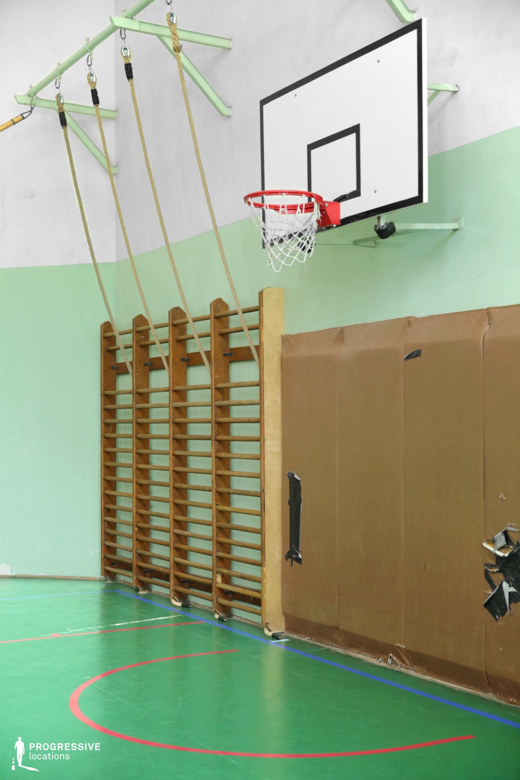 This retro-style gymnasium corner features a mounted basketball hoop above a padded wall, wooden wall bars, and hanging climbing ropes, evoking a classic school sports setting. The green and white walls contrast with the polished green sports floor, while the aged protective padding adds an authentic, slightly worn aesthetic. The utilitarian lighting and exposed metal brackets contribute to an old-school athletic environment. This location is well-suited for filming sports dramas, nostalgic school scenes, or training montages, offering a raw, functional backdrop ideal for themes of perseverance, rivalry, or discipline.