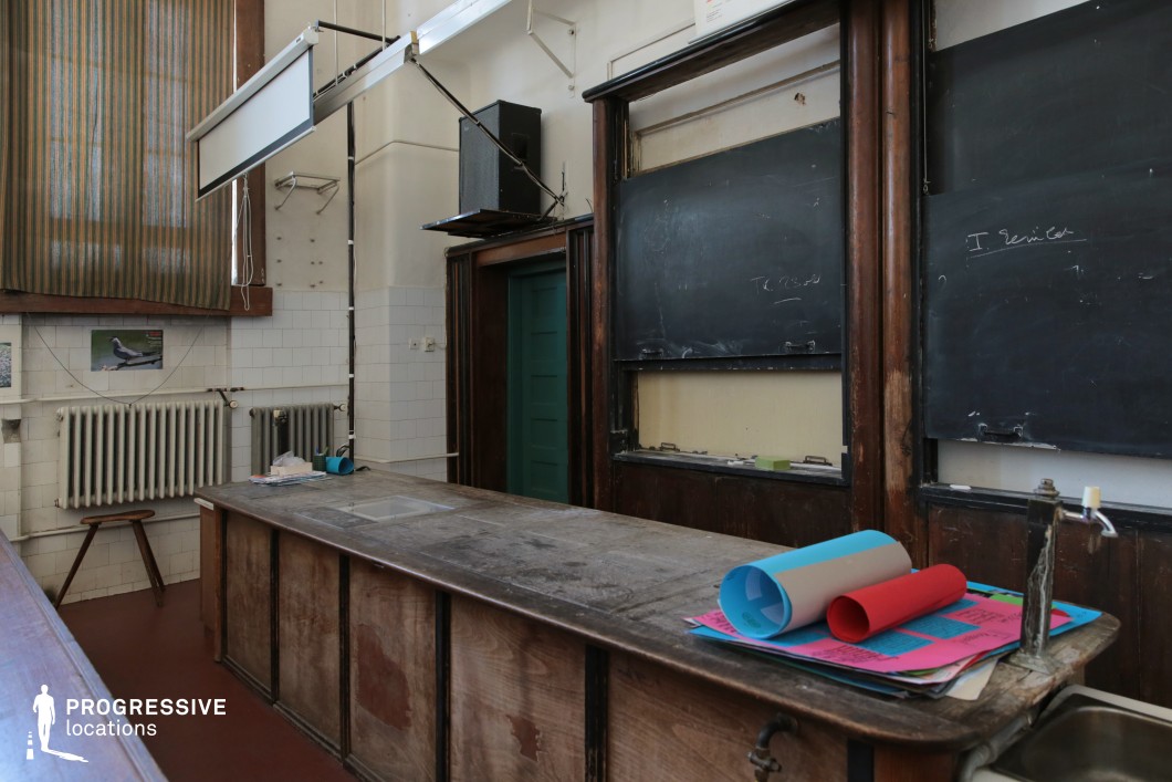 This retro-style classroom features dark wooden paneling, aged blackboards, and a worn teacher’s desk, evoking an old-fashioned academic atmosphere. The white tiled walls contrast with the deep tones of the wood, while a green door and vintage radiator add to the historical charm. The overhead projector screen and scattered teaching materials suggest a well-used educational space with a layered history. Ideal for period dramas, academic thrillers, or nostalgic coming-of-age films, this setting provides an authentic backdrop for narratives centered around knowledge, authority, or intellectual discovery.