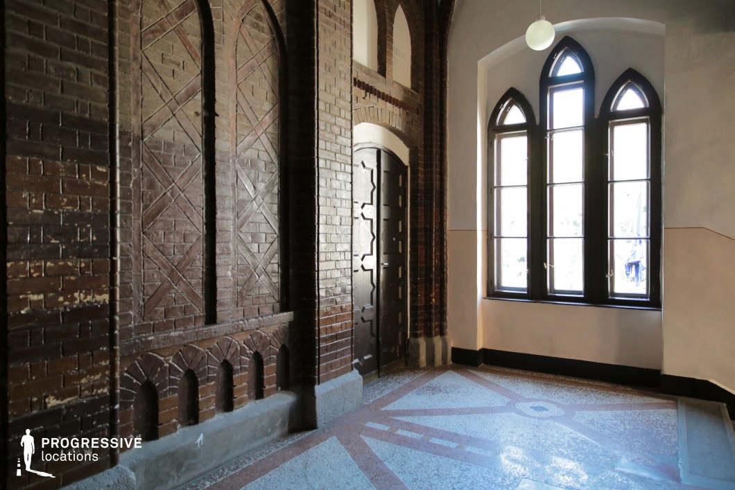 This historic interior space features a rich blend of Gothic and industrial elements, with dark exposed brick walls showcasing decorative archways and intricate geometric patterns. The large, pointed-arch windows framed in black wood allow natural light to filter in, enhancing the moody yet elegant atmosphere. The terrazzo flooring with ornamental inlays adds a refined contrast to the raw textures of the brick. Ideal for period dramas, mystery thrillers, or historical fiction, this setting evokes a sense of tradition, secrecy, and timeless grandeur.