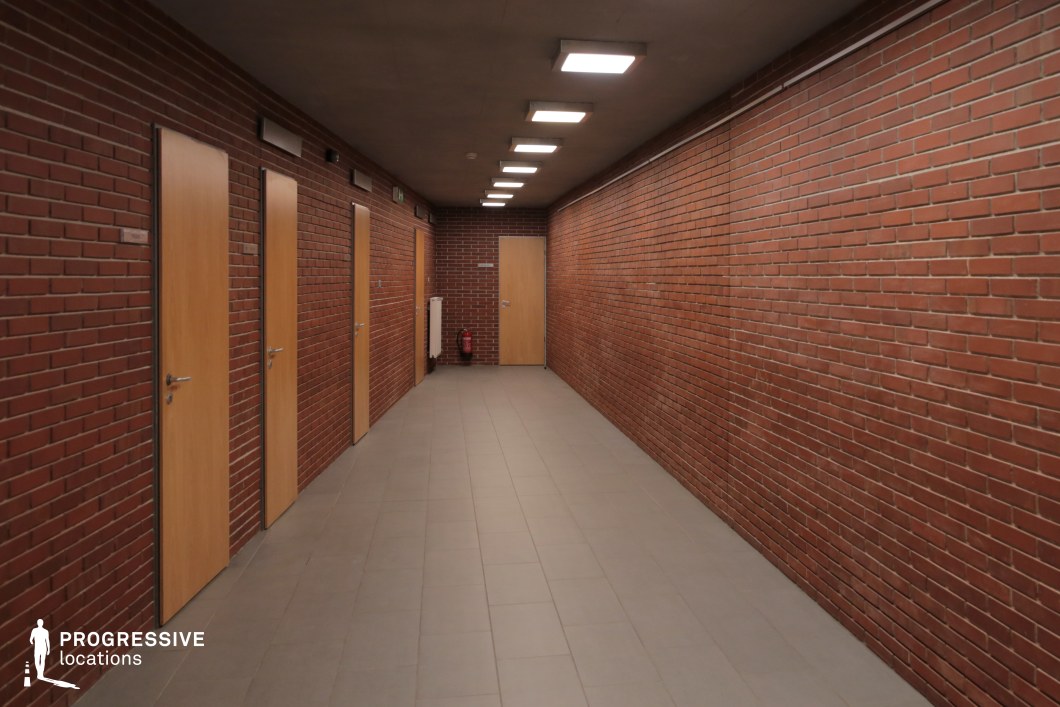 This enclosed corridor features exposed red brick walls, evenly spaced wooden doors, and a tiled floor, creating a structured and utilitarian aesthetic. The low ceiling, fitted with rectangular recessed lighting, casts a uniform glow throughout the space, enhancing the sense of depth. The combination of warm wood tones and industrial brick textures gives the space a functional yet slightly rigid atmosphere. This location is well-suited for suspenseful, institutional, or academic settings, making it ideal for thriller, drama, or investigative film genres requiring a controlled and somewhat enclosed environment.