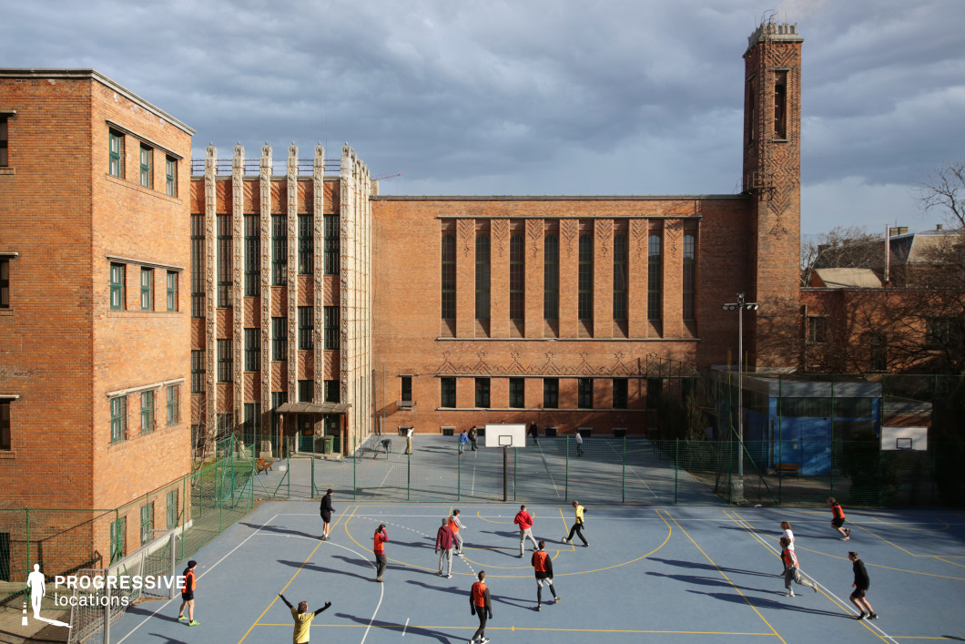 A striking Art Deco-inspired brick building dominates the scene, with tall, narrow windows and intricate geometric detailing. The courtyard features a blue outdoor sports court, enclosed by a green metal fence. The imposing structure includes a tall, narrow chimney, adding to its historical character. The setting is ideal for academic or institutional backdrops, sports-related productions, or period dramas requiring an early 20th-century urban environment. The open courtyard provides ample space for dynamic action sequences or atmospheric shots of school life.