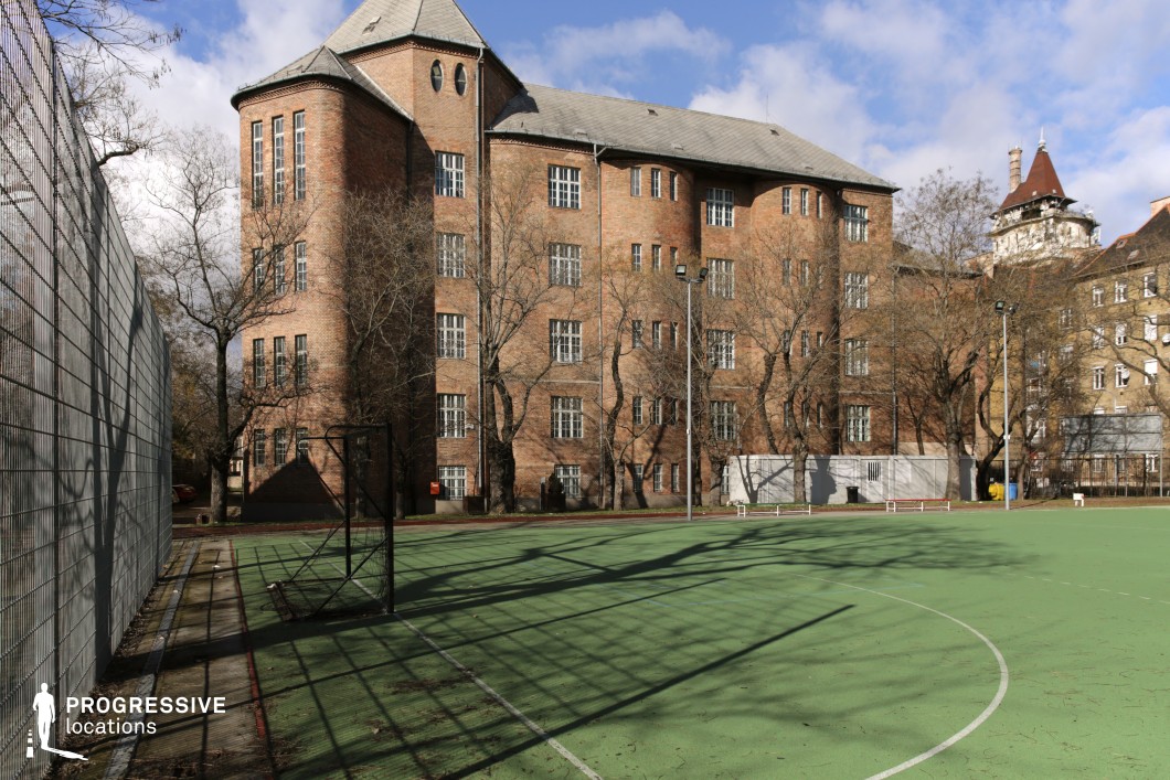 This location features a striking multi-story brick building with a historic architectural style, characterized by arched and rectangular windows, steeply pitched roofs, and a tower-like structure. The warm-toned brick facade is complemented by leafless trees, adding a stark contrast to the rigid geometry of the structure. In the foreground, a fenced sports court with a green playing surface extends into the frame, bordered by metal mesh fencing and tall floodlights. The combination of the imposing institutional-style architecture and the open recreational area creates a visually compelling contrast. This location is ideal for filming school, institutional, or historical drama scenes, as well as suspense or mystery genres requiring a secluded or atmospheric backdrop.