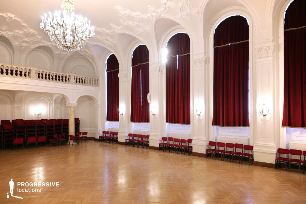 This opulent ceremonial hall features soaring arched windows framed by deep red velvet curtains, intricate white stucco moldings, and a grand chandelier that illuminates the polished parquet floor. The high ceilings and decorative balcony add to the room’s sense of grandeur, making it an ideal setting for elegant gatherings, formal events, or historical reenactments. The symmetry, rich textures, and classical architectural elements create a visually striking space, perfect for period dramas, ballroom scenes, aristocratic settings, or films exploring themes of power, tradition, and prestige.