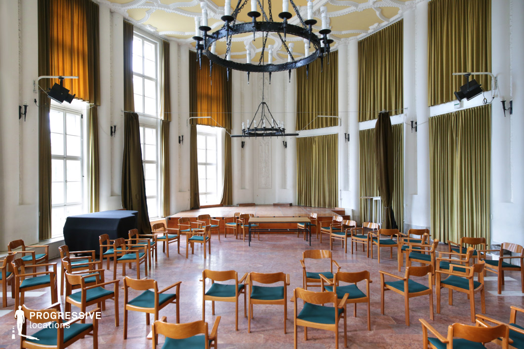 A spacious ceremonial hall featuring a vintage aesthetic with high ceilings, tall arched windows, and ornate molding details. The room is illuminated by natural light filtering through the large windows, adorned with heavy olive-green curtains that add warmth to the white walls. Suspended from the intricately decorated ceiling are wrought-iron chandeliers with candle-like fixtures, reinforcing the historic ambiance. The polished wooden floor complements the retro-style wooden chairs with dark green upholstery, arranged in a scattered formation. The elevated stage area, framed by curtains, suggests a setting suitable for performances, lectures, or formal events. This location offers a refined and timeless atmosphere, ideal for period films, artistic performances, or sophisticated academic scenes.