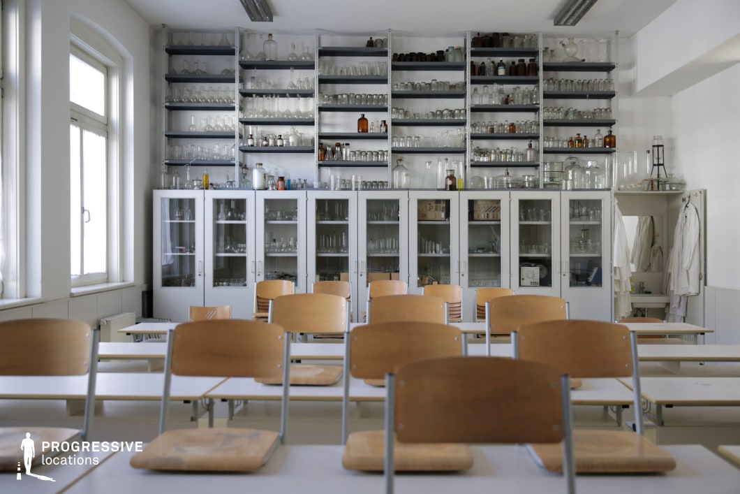 This well-preserved chemistry classroom features a structured layout with wooden chairs and white tabletops arranged in rows facing a wall of cabinets filled with an extensive collection of laboratory glassware. The tall shelves, housing beakers, flasks, and reagent bottles, contribute to the room’s academic and scientific character. Natural light streams in through large arched windows, enhancing the clean, white aesthetic and highlighting the practical yet historical ambiance of the space. White lab coats hang by a small sink area, adding to the authenticity of a working laboratory setting. This location is ideal for period academic scenes, science-based storytelling, or nostalgic educational visuals.