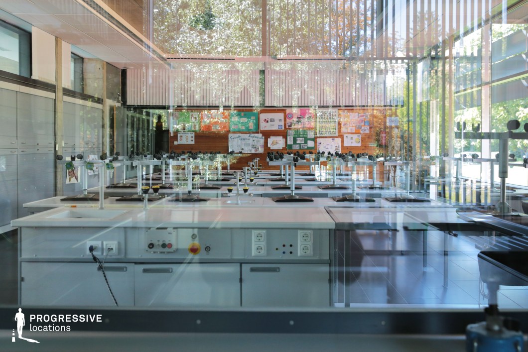 This contemporary science laboratory features a sleek, functional design with clean white workstations, built-in gas taps, and an array of electrical outlets for experiments. The space is enclosed by floor-to-ceiling glass walls, allowing natural light to blend with reflections of the surrounding greenery. The interior combines industrial and warm wooden elements, with educational posters adorning the back wall, adding a scholastic touch. The structured symmetry of the workstations enhances the futuristic aesthetic, making this an ideal filming location for academic, sci-fi, or investigative drama genres, particularly scenes involving research, innovation, or discovery.