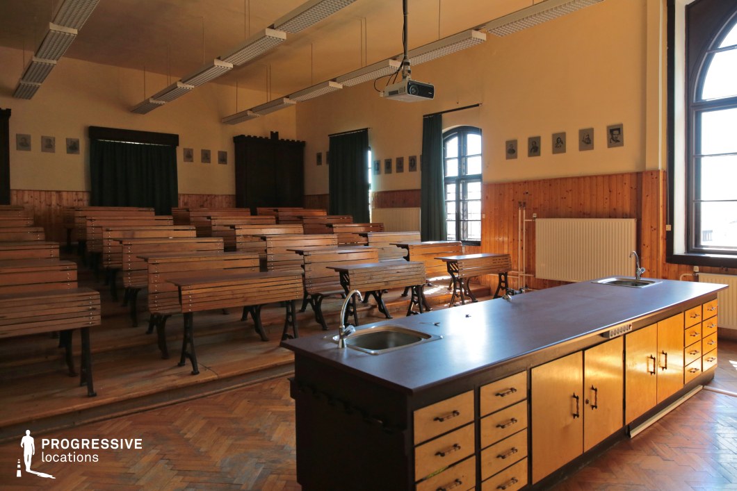 This traditional lecture hall features tiered wooden benches with cast-iron legs, exuding a historical academic atmosphere. The room is lined with warm wooden paneling, contrasted by cream-colored walls adorned with framed portraits. Large arched windows allow natural light to cast a soft glow on the parquet flooring and laboratory-style demonstration desk equipped with sinks. Overhead, a suspended projector modernizes the otherwise vintage setting. Ideal for period dramas, academic thrillers, or coming-of-age films, this space evokes themes of knowledge, discipline, and intellectual pursuit.