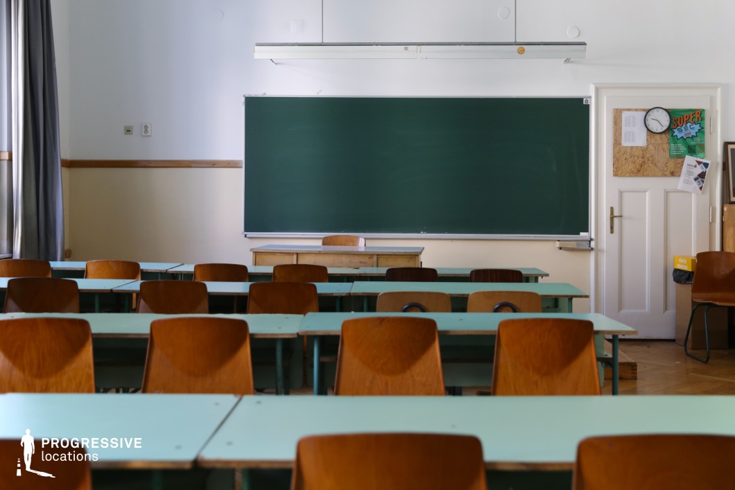 This vintage-style classroom features rows of mint-green desks and warm wooden chairs, all facing a large green chalkboard. The walls are painted in a muted off-white with a wooden trim, contributing to the structured academic ambiance. A teacher’s wooden desk sits in front of the board, reinforcing the classic school setting. The large windows with gray curtains allow soft, natural light to filter in, creating a well-lit yet slightly aged atmosphere. Ideal for filming school-related dramas, coming-of-age films, or historical settings, this location offers an authentic educational backdrop with a timeless, institutional feel.