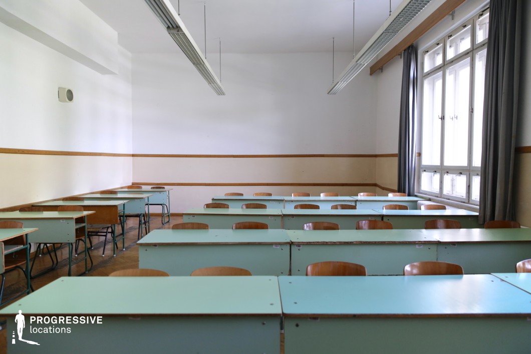 This vintage-style classroom is characterized by rows of pale green desks and wooden chairs, arranged in a structured layout that evokes a traditional academic setting. The neutral off-white walls with a horizontal wooden trim add to the subdued, institutional aesthetic. Large windows with gray curtains allow soft, natural light to filter in, while overhead fluorescent tube lights reinforce the retro atmosphere. The aged wooden parquet flooring contrasts with the utilitarian furnishings. Ideal for filming school-related dramas, coming-of-age stories, or historical settings, this location provides a timeless educational backdrop suitable for both nostalgic and introspective narratives.
