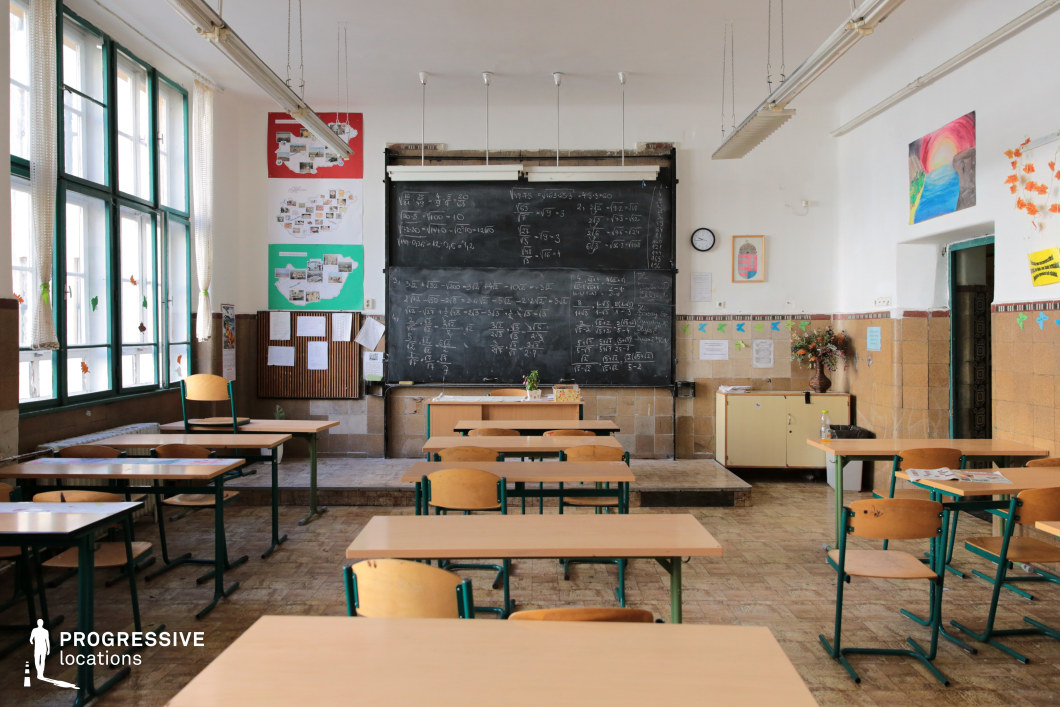 This classroom features a vintage academic setting with a large, well-worn blackboard covered in mathematical equations as the focal point. The space has a warm, aged atmosphere, with beige tiled walls, wooden furniture, and green-framed windows that let in abundant natural light. The neatly arranged desks and chairs contrast with the slightly worn floor, while educational posters and student artwork add personality to the environment. Suspended fluorescent lighting and high ceilings contribute to the institutional character. This location is ideal for school-related films, period dramas, or coming-of-age stories, offering an authentic and timeless classroom aesthetic.