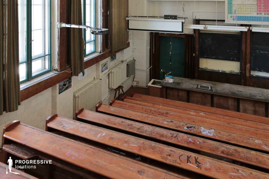 This aged lecture hall features tiered wooden benches, deeply worn and covered in carved markings, evoking a sense of history and student culture. The front of the room is dominated by a long, well-used wooden demonstration table, flanked by dark paneling and old blackboards. Large windows with blue frames and striped curtains filter in natural light, while tiled walls and vintage radiators reinforce the academic atmosphere. The mix of aged materials and functional design makes this location ideal for period dramas, coming-of-age stories, or narratives exploring themes of knowledge, rebellion, or nostalgia in an institutional setting.