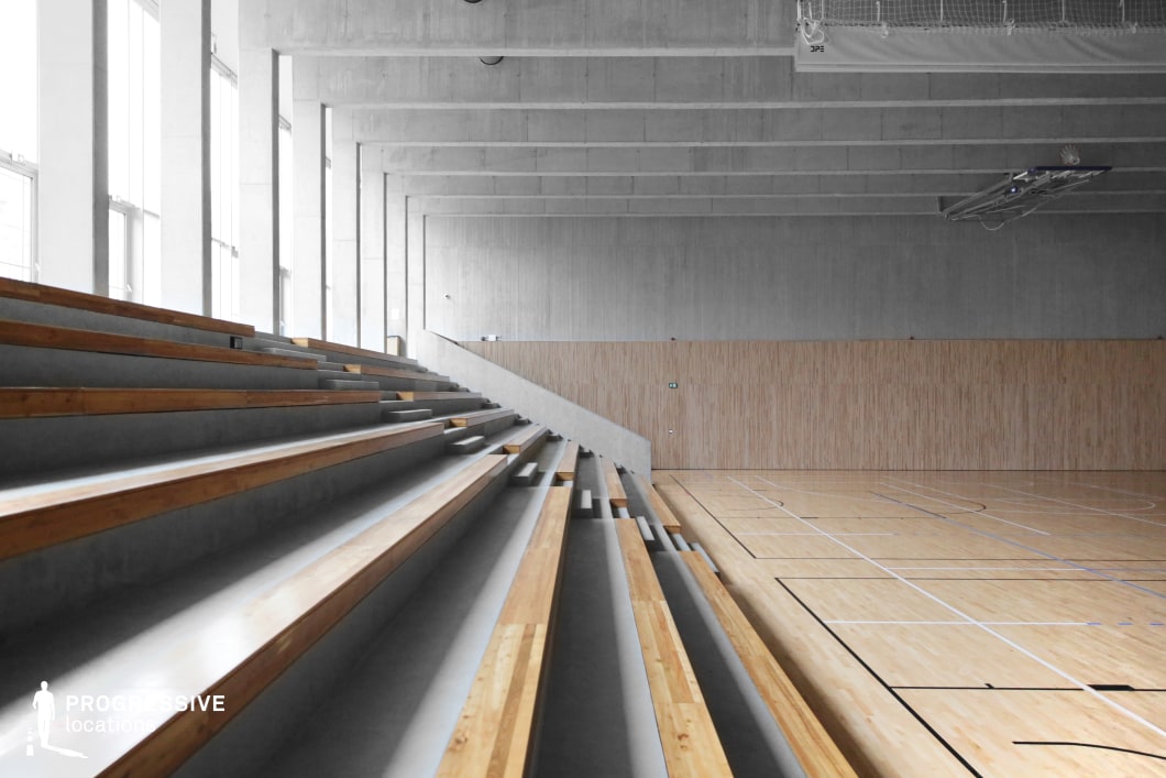 This modern sports hall features a sleek, minimalist design with raw concrete surfaces, wooden paneling, and clean architectural lines. The bleachers, made of smooth concrete with wooden seating strips, extend alongside the polished wooden court marked for multiple sports. Large floor-to-ceiling windows allow natural light to illuminate the space, enhancing its open and airy feel. The combination of industrial materials and warm wood creates a balanced aesthetic that is both contemporary and functional. This location is ideal for filming sports dramas, training montages, or atmospheric scenes in dystopian, indie, or architectural-focused productions.