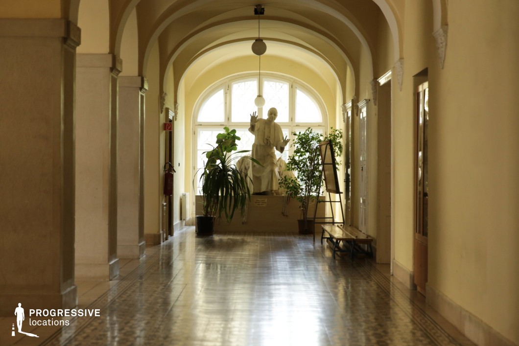 This classical corridor exudes historical and academic charm, featuring a series of soft yellow-toned arches supported by white stone columns. The polished patterned tile flooring reflects natural light streaming from an arched window at the far end, where a sculpted statue is prominently displayed, framed by lush green plants. Wooden and glass-paneled doors line the hallway, enhancing the vintage character, while intricate decorative moldings and suspended spherical light fixtures contribute to the refined ambiance. The corridor’s symmetry and depth make it a visually striking filming location, ideal for period dramas, mystery thrillers, or historical documentaries requiring an elegant and timeless backdrop.