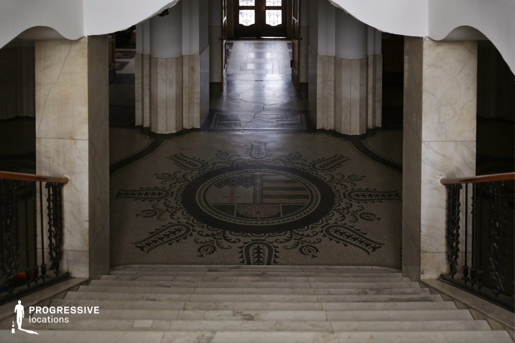 This stately staircase descends into an elegant hall, featuring a detailed mosaic floor with a heraldic design at its center. The surrounding architecture is characterized by marble-clad columns, intricate wrought-iron railings, and smooth white arches, creating a refined yet slightly aged atmosphere. The interplay of shadow and light enhances the depth of the scene, with a set of double doors in the background suggesting an imposing entrance. This location is well-suited for historical dramas, mystery thrillers, or institutional settings, offering a sense of grandeur, authority, and intrigue.
