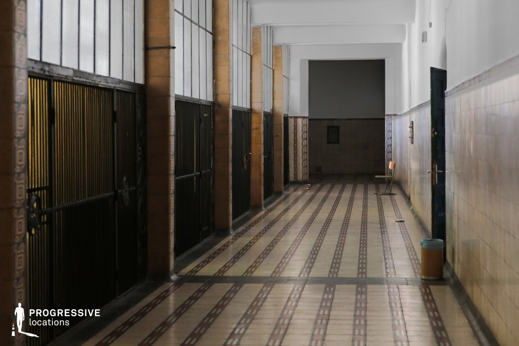 This narrow, tiled corridor exudes a sense of vintage institutional architecture, possibly reminiscent of an old school, archive, or even a former detention area. The alternating black and beige patterned tiles add depth to the scene, leading the eye towards a darkened passage at the far end. Tall, iron-barred doors line one side, framed by warm-toned stone columns and frosted glass windows above, allowing diffused light to enter. The opposite side features tiled walls, with a single chair subtly emphasizing the emptiness of the space. Ideal for scenes evoking secrecy, nostalgia, or confinement, this location lends itself well to historical dramas, suspenseful narratives, or institutional settings.