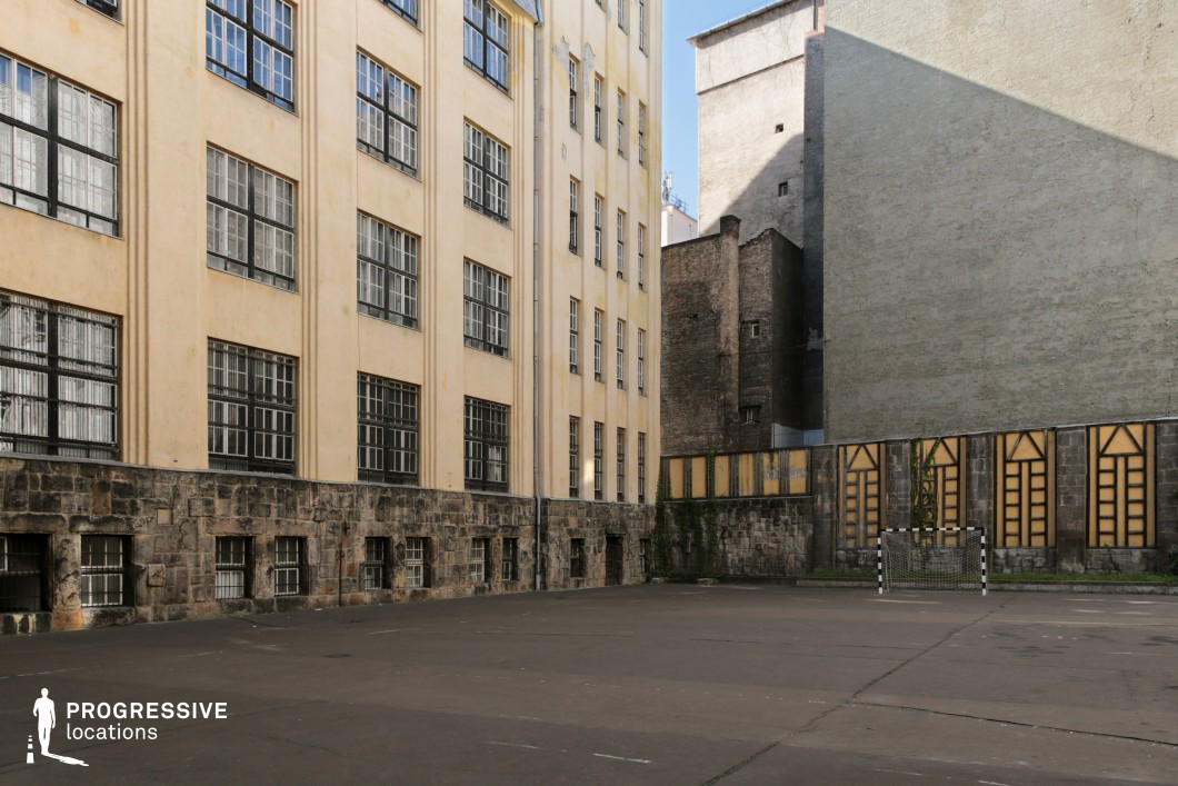 This enclosed courtyard, framed by tall, historic buildings with a mix of stone and plaster facades, exudes a raw, institutional character. The worn asphalt surface, barred windows, and a single goalpost contribute to its utilitarian, slightly austere atmosphere. The surrounding aged brick walls and geometric-patterned fencing add texture, while the contrast between the yellowed facades and gray concrete structures enhances the urban aesthetic. Ideal for gritty dramas, coming-of-age films, or historical narratives, this location offers a versatile setting for themes of isolation, nostalgia, or resilience.